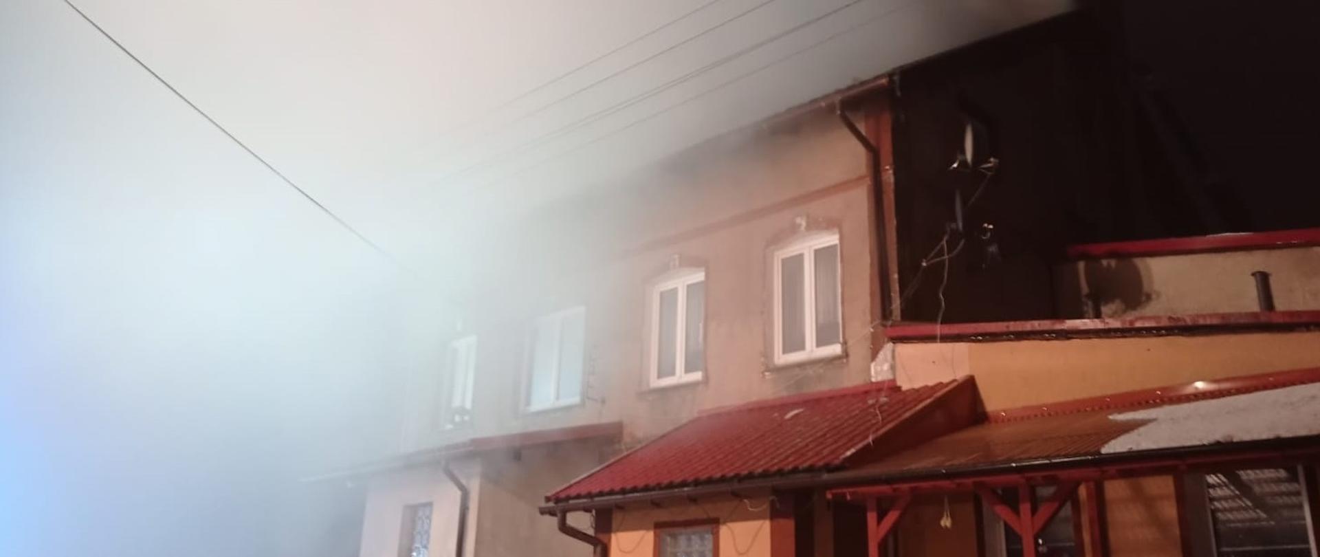 Zadymiony budynek wielorodzinny, dym wydobywa się z dachu. Przed budynkiem stoi dwóch strażaków ubranych w ubranie bojowe. Strażacy zabezpieczają linię gaśniczą. 