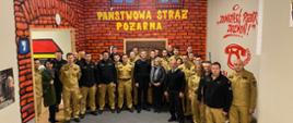 Pomieszczenie Centrum Edukacji Przeciwpożarowej. Na ścianach namalowane cegły, okna i napis Państwowa Straż Pożarna. Na tle ściany uczestnicy szkolenia pozują do grupowego zdjęcia. 