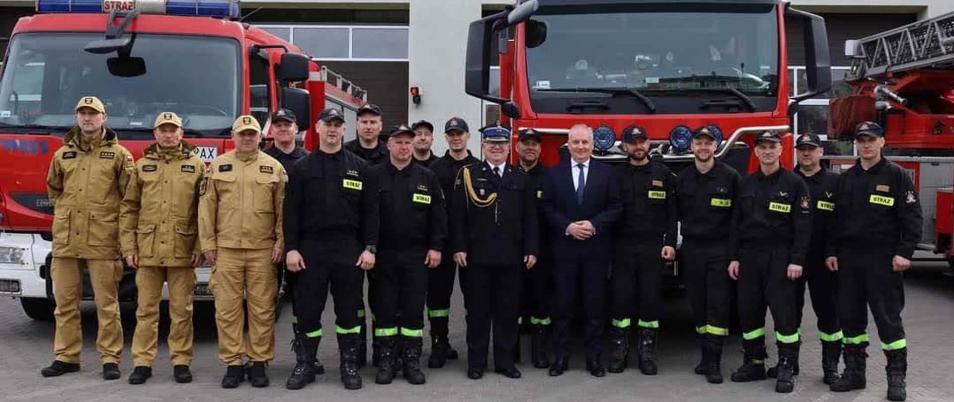Strażacy oraz mężczyzna w garniturze stoją obok siebie za nimi ustawione są samochody gaśnicze.