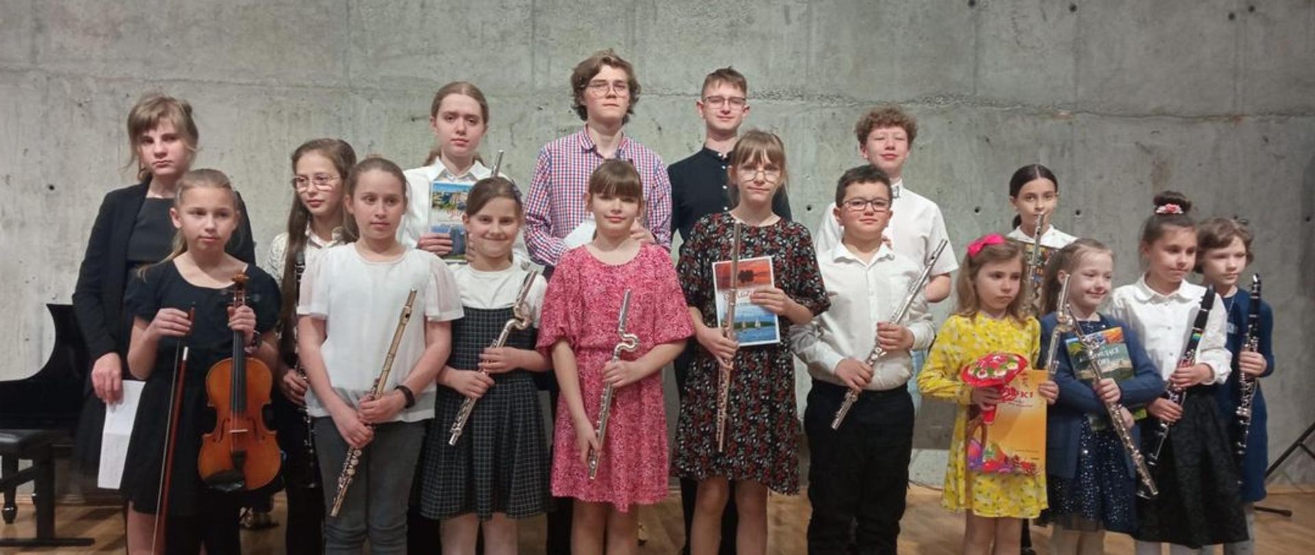 Grupa uczniów trzymających w rękach instrumenty stoi na scenie sali koncertowej szkoły muzycznej