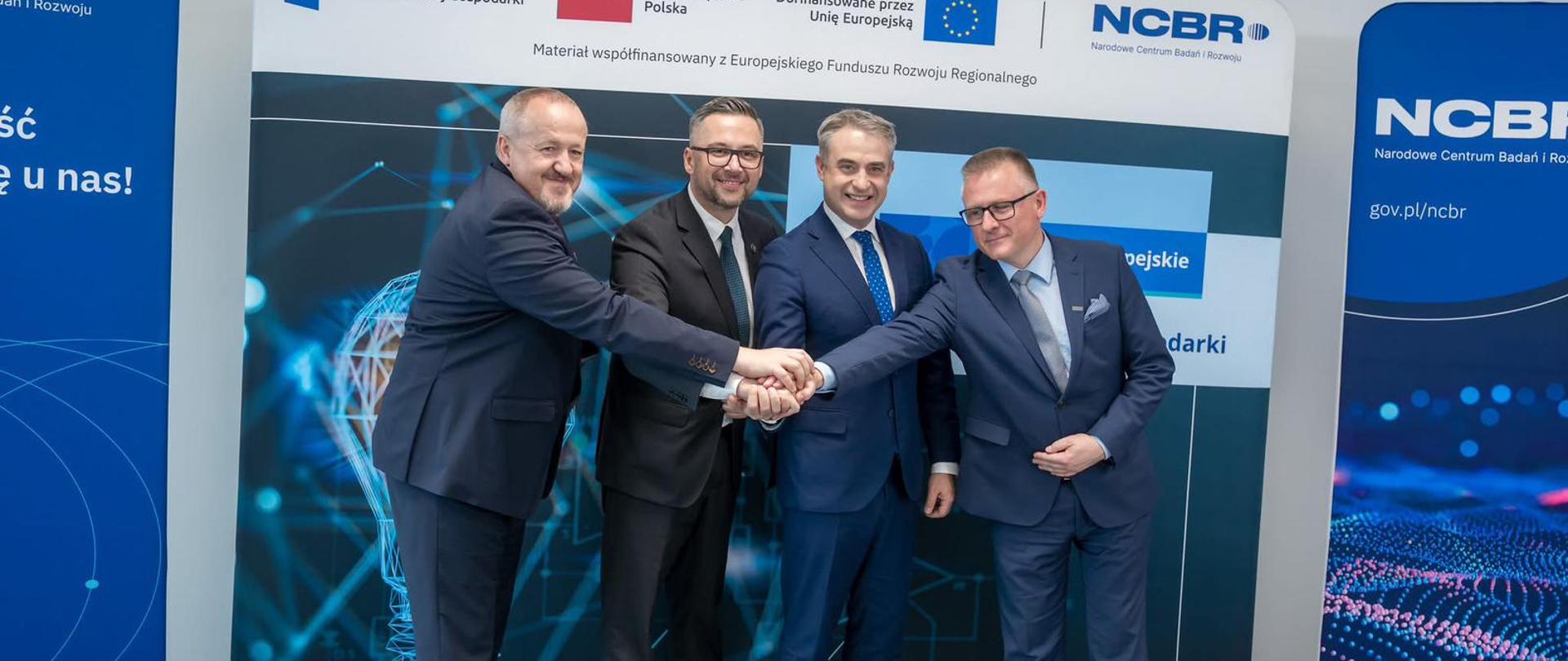 Na tle banerów z napisem NCBR stoi czterech mężczyzn, wszyscy trzymają się za ręce, drugi od lewej minister Kulasek.
