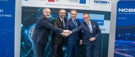 Na tle banerów z napisem NCBR stoi czterech mężczyzn, wszyscy trzymają się za ręce, drugi od lewej minister Kulasek.
