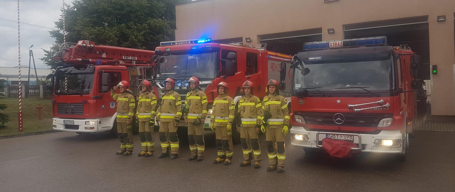 Na zdjęciu widać strażaków w umundurowaniu bojowym ustawionych w szyku na tle samochodów pożarniczych.