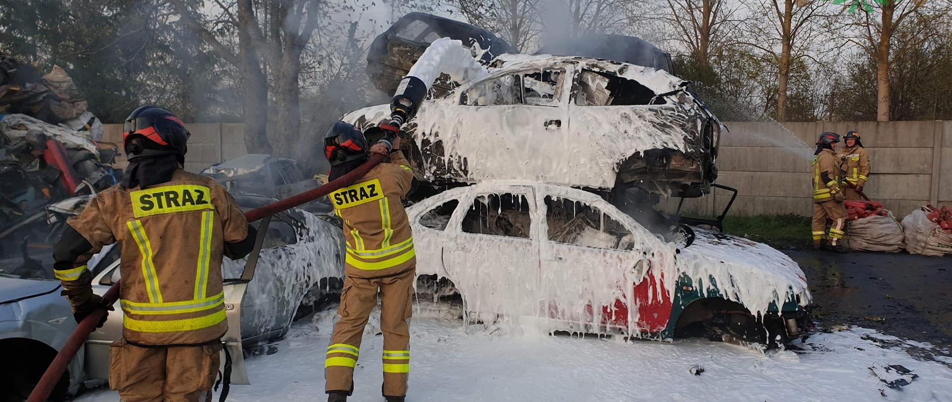 Strażacy podają prąd piany na wraki samochodów ułożonych jeden na drugim.