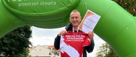 Zdjęcie przedstawia mężczyznę trzymającego koszulkę z napisem Mistrz Polski Strażaków oraz dyplom. W tle meta zawodów Dycha Przemyska
