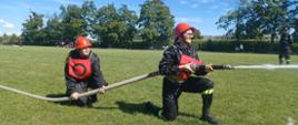 Na zdjęciu widać dwie dziewczyny w czarnym stroju strażackim. Na głowie mają czerwone hełmy. Jedna z zawodniczek trzyma prądownice i leje wodą. Druga, która znajduje się za nią trzyma wąż. Konkurencja odbywa się na trawie. W tle widać sędziego zawodów, grupy kibiców, budynki, drzewa oraz kukurydze rosnąca na polu. Jest widno.