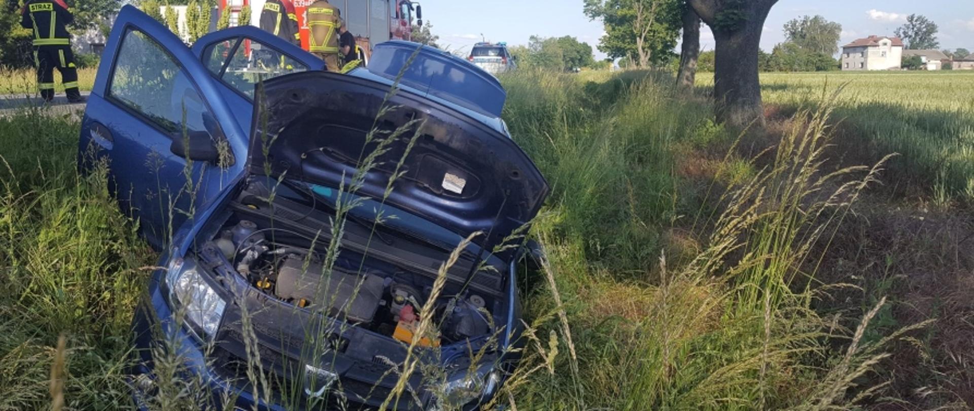 Na zdjęciu widoczny samochód osobowy koloru niebieskiego w przydrożnym rowie. Na drugim planie na jezdni widoczny radiowóz oraz samochód straży pożarnej.