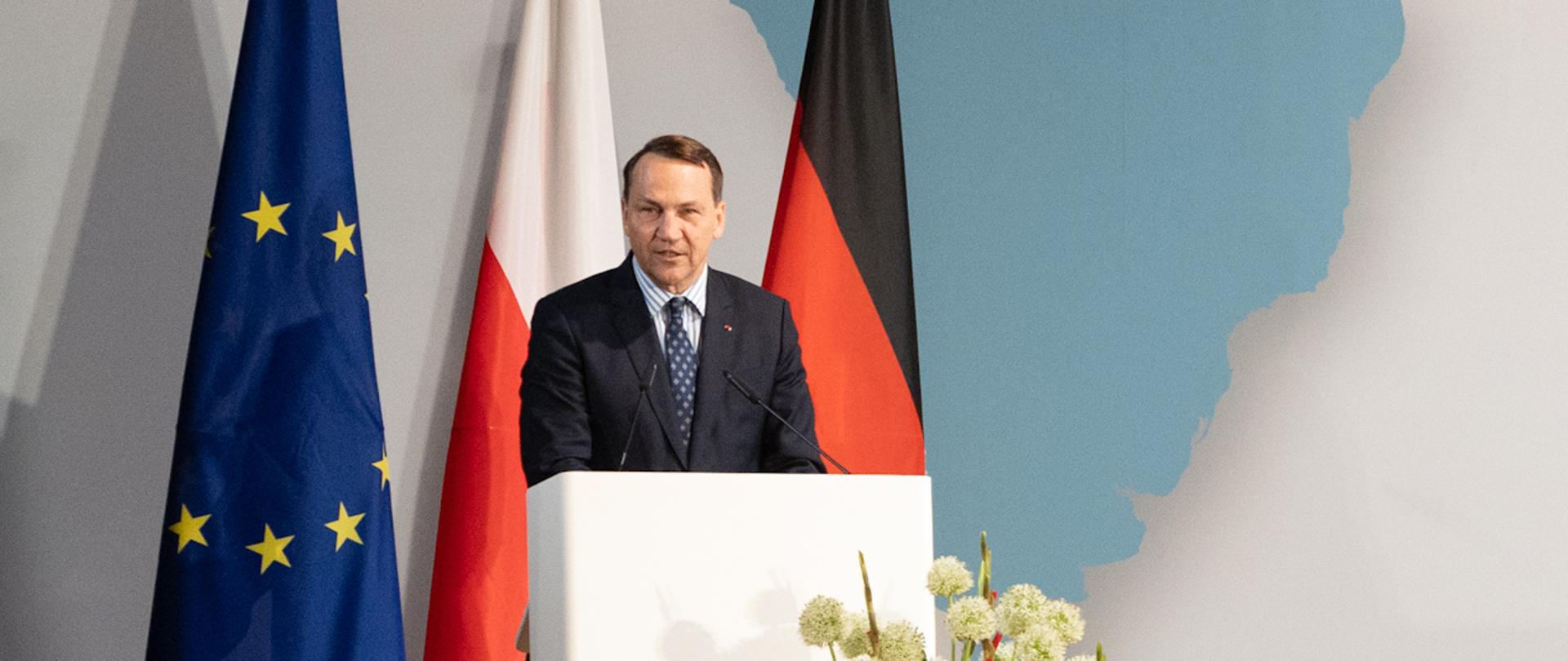 Wicepremier Sikorski w granatowym garniturze i niebieskim krawacie przemawia z mównicy podczas Polsko-Niemieckiego Forum w Berlinie. Za nim stoją flagi Unii Europejskiej, Polski i Niemiec oraz duża ściana z mapą świata i napisem „Deutsch-Polnisches Forum / Polsko-Niemieckie Forum, Berlin, 17.06.2026”. Po prawej stronie sceny tłumacz języka migowego w ciemnym stroju przekłada wystąpienie. Na widowni zgromadzona jest liczna publiczność siedząca w rzędach, część uczestników fotografuje wydarzenie telefonami.