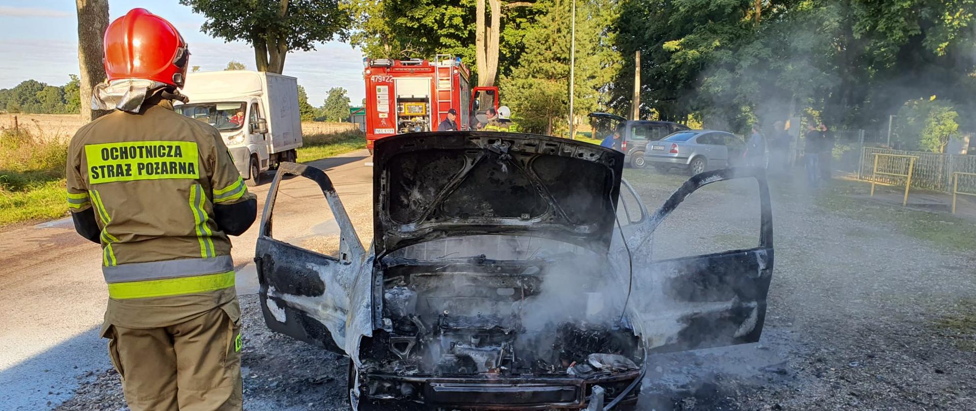 Zdjęcie przedstawia miejsce zdarzenia- spalony samochód osobowy oraz strażaka ubranego w piaskowe ubranie specjalne oraz czerwony hełm. W tle inne samochody oraz czerwony wóz bojowy.