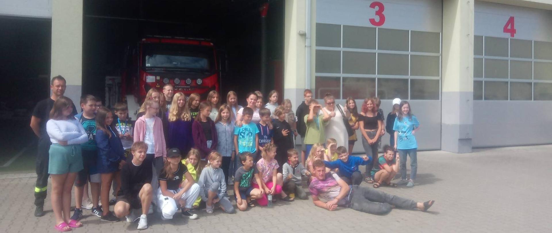 Wycieczka uczestników kolonii do KP PSP w Sokołowie Podlaskim - na zdjęciu strażacy i dzieci z kolonii na placu przed strażnicą na zbiórce przed garażem.