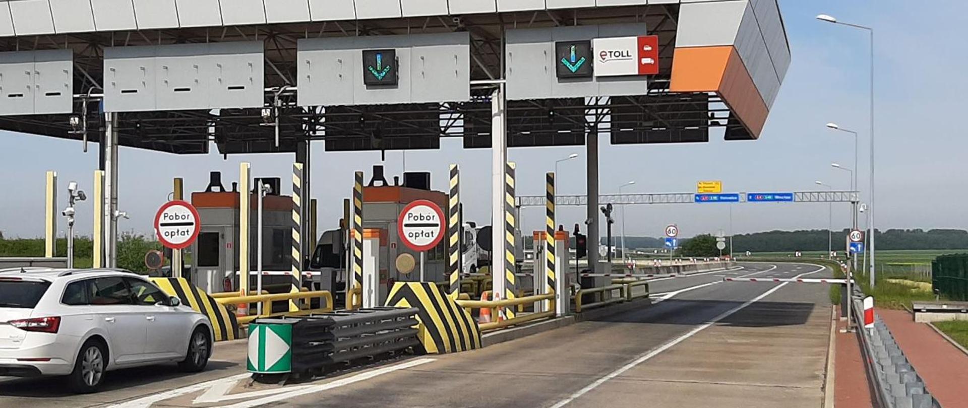 Bramki poboru opłat e‑TOLL na drodze szybkiego ruchu. Nad pasami ruchu wiszą sygnalizatory z zielonymi strzałkami. Przy bramkach stoją znaki „Pobór opłat”. Po lewej stronie widać samochód osobowy, w tle otwarta droga i znaki drogowe.