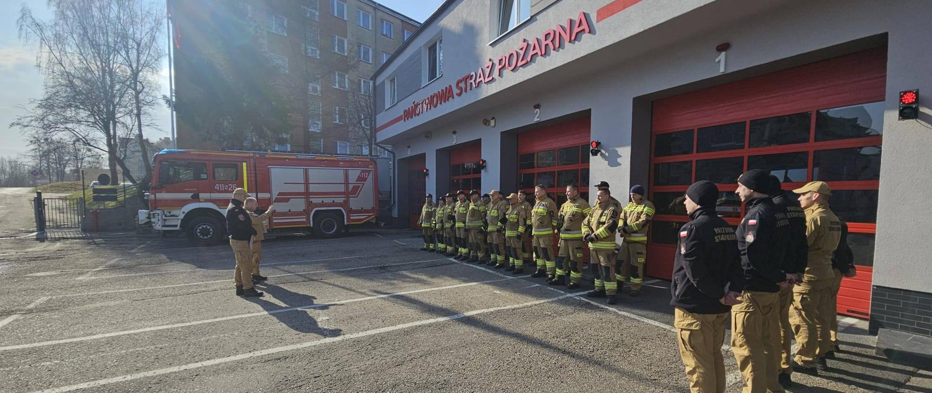 Plac wewnętrzny jednostki ratowniczo gaśniczej. Z prawej strony budynek remizy z czerwonymi dużymi bramami. Przy budynku w dwuszeregu strażacy w mundurach bojowych. Słonecznie.