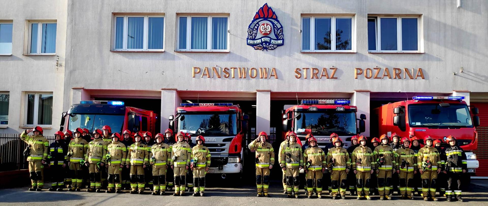 Zdjęcie przedstawia stojących w dwuszeregu strażaków w umundurowaniu specjalnym koloru piaskowego. Komendy Miejskiej PSP na tle ściany frontowej budynku komendy. Ba ścianie powyżej, pomiędzy oknami znajduje się znak graficzny Państwowej Straży Pożarnej oraz napis Państwowa Straż Pożarna. Za strażakami widoczne są cztery samochody pożarnicze
