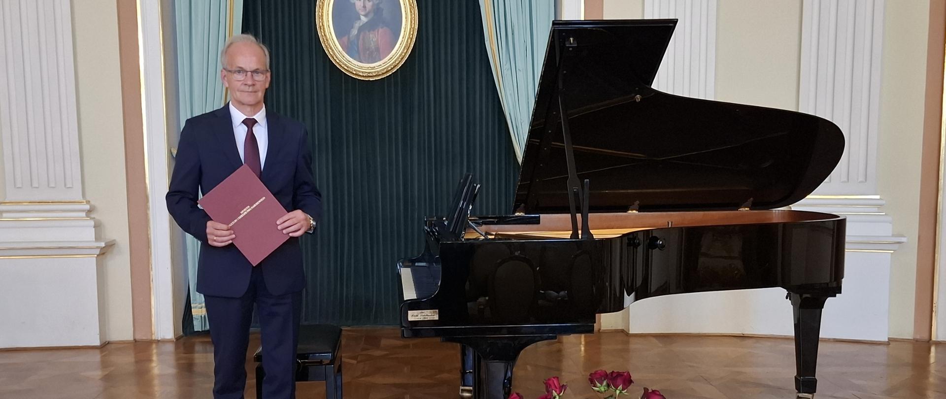 Mężczyzna ubrany w granatowy garnitur stoi i pozuje do zdjęcia. Z jego lewej strony stoi czarny fortepian. Przed fortepianem na podłodze stoi wazon z czerwonymi różami.