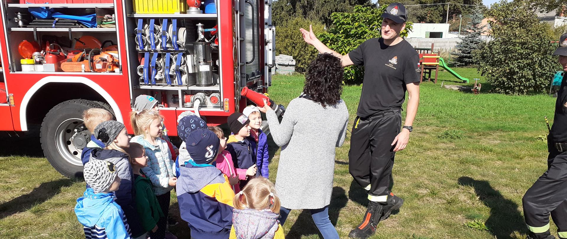 Na zdjęciu widać grupę przedszkolaków, biorących udział w pokazie prowadzonym przez strażaków. Dzieci przyglądają się strażakowi, który prowadzi pogadankę. Opiekunka stoi w grupie dzieci i trzyma prądownicę. W tle widać samochód ratowniczo-gaśniczy z otwartymi skrytkami. Całość odbywa się na trawie na boisku przy budynku przedszkola.