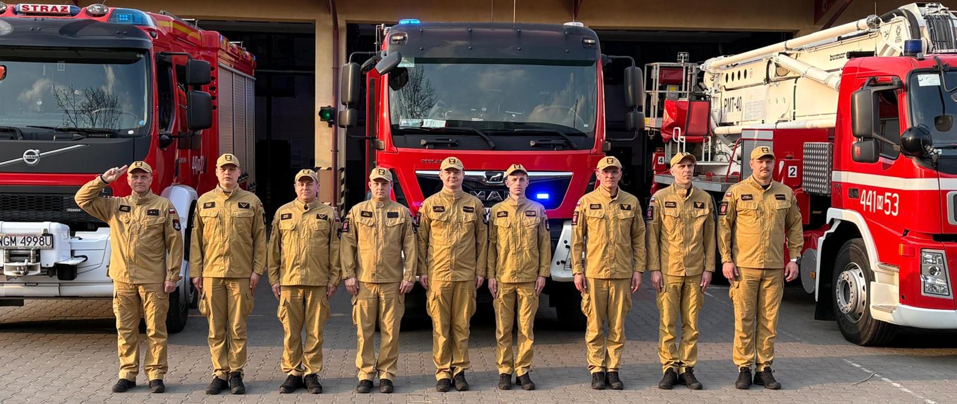 Dziewięciu strażaków w piaskowych mundurach służbowych stoi w równym szeregu na placu przed remizą. Pierwszy mężczyzna z lewej salutuje, podczas gdy pozostali zachowują postawę zasadniczą. W tle zaparkowane są trzy czerwone wozy strażackie marek Volvo i MAN oraz pojazd z podnośnikiem hydraulicznym. Scena rozgrywa się w słoneczny dzień na wybrukowanym podjeździe jednostki straży pożarnej.