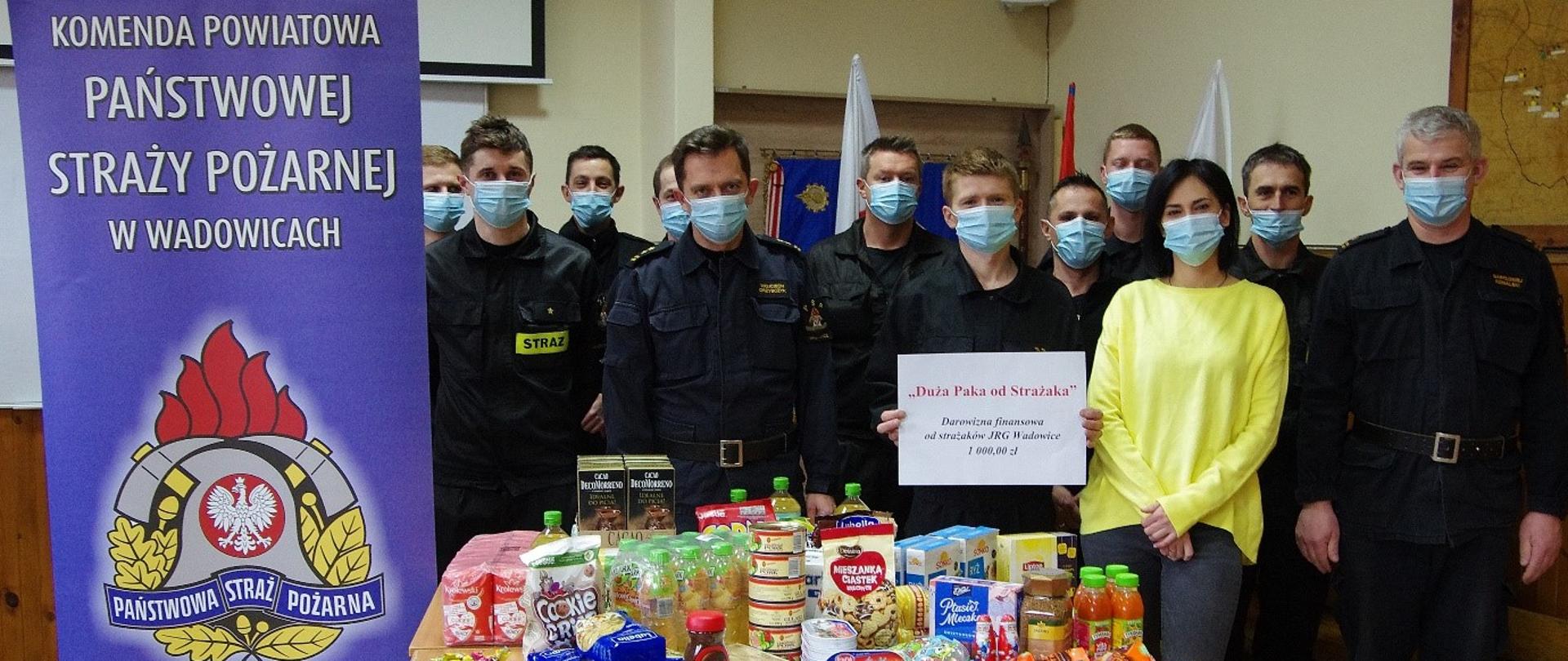 Grupa strażaków w umundurowaniu koszarowym w maseczkach. Przed nimi produkty spożywcze ustawione na stole. Jeden ze strażaków trzyma kartkę z napisem Duża Paka od Strażaka Darowizna finansowa od strażaków JRG Wadowice 1000 zł. Po lewej stronie widoczny rollup JRG w Wadowicach z logo PSP.