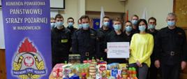Grupa strażaków w umundurowaniu koszarowym w maseczkach. Przed nimi produkty spożywcze ustawione na stole. Jeden ze strażaków trzyma kartkę z napisem Duża Paka od Strażaka Darowizna finansowa od strażaków JRG Wadowice 1000 zł. Po lewej stronie widoczny rollup JRG w Wadowicach z logo PSP.