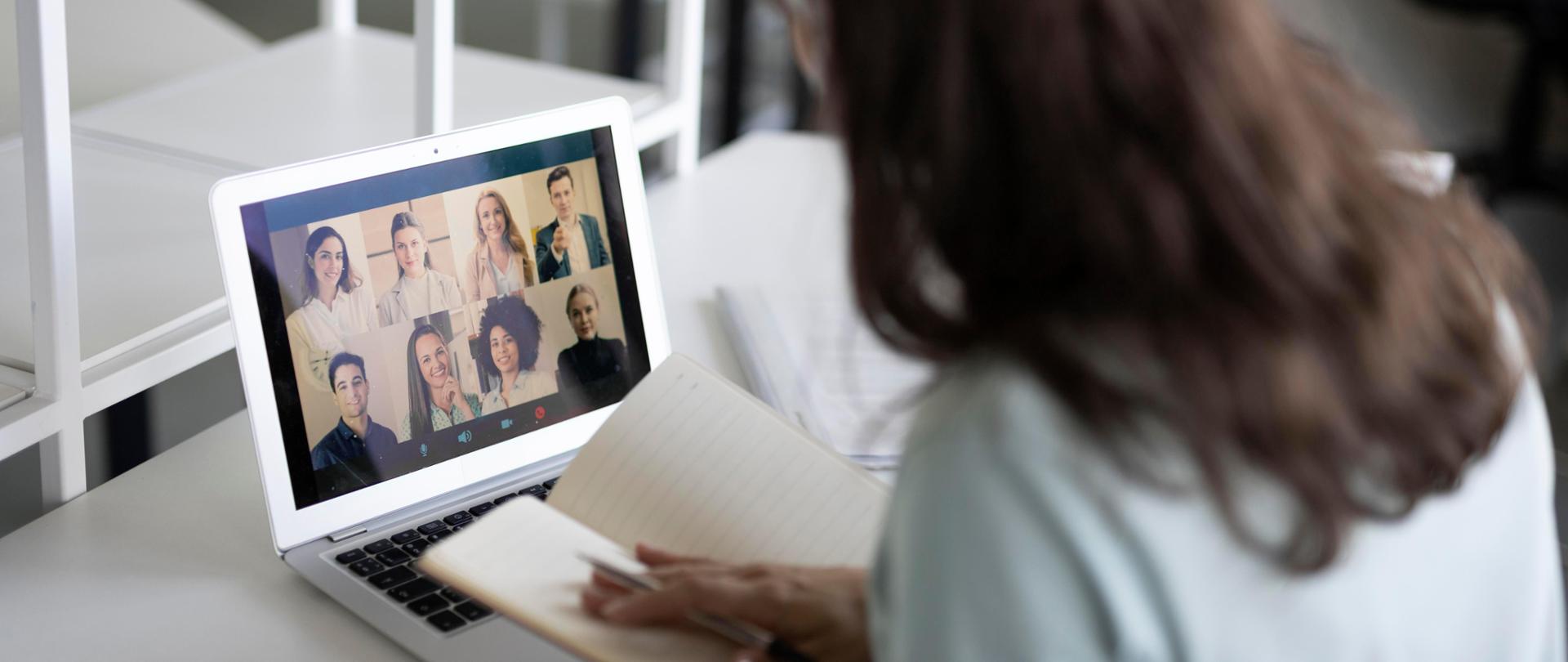 Zdjęcie z tyłu kobiety z notatnikiem, spoglądającej na komputer, na którym jest prowadzona wideokonferencja. 