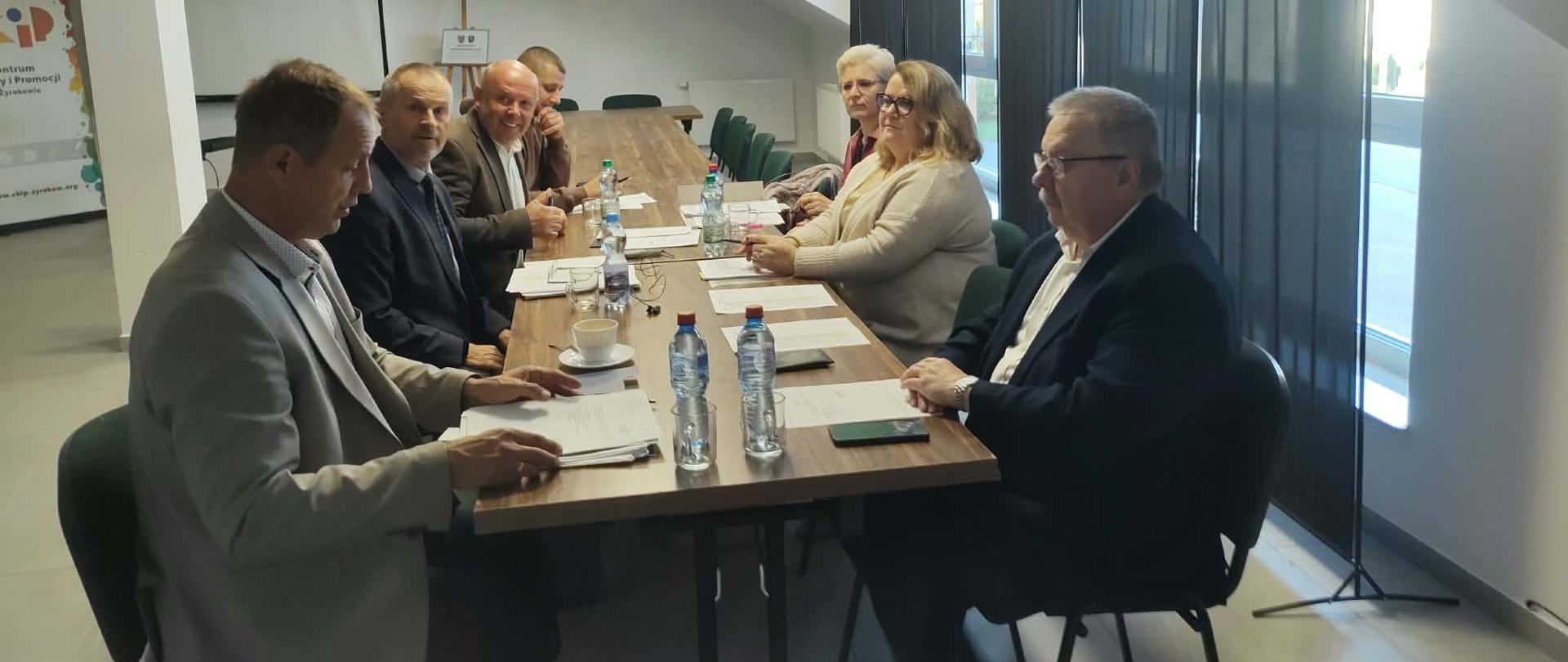 Na zdjęciu widać grupę siedmiu osób podczas spotkania przy długim, drewnianym stole w jasnej sali konferencyjnej. Uczestnicy siedzą po obu stronach stołu, na którym znajdują się dokumenty, butelki i szklanki z wodą, filiżanka kawy oraz telefony.
Na pierwszym planie, po lewej stronie, siedzi mężczyzna w jasnej marynarce Piotr Giza - Kierownik Wojewódzkiego Inspektoratu Ochrony Roślin i Nasiennictwa w Rzeszowie, Oddział w Dębicy, pochylony nad dokumentami. Po jego lewej stronie siedzi kilku mężczyzn w marynarkach. Po przeciwnej stronie stołu znajdują się dwie kobiety oraz mężczyzna w ciemnej marynarce Wójt Gminy Żyraków, który siedzi najbliżej prawej krawędzi zdjęcia.
W tle znajdują się duże okna. Okna są zasłonięte ciemnymi, składanymi panelami. Nad stołem znajdują się prostokątne lampy sufitowe.
