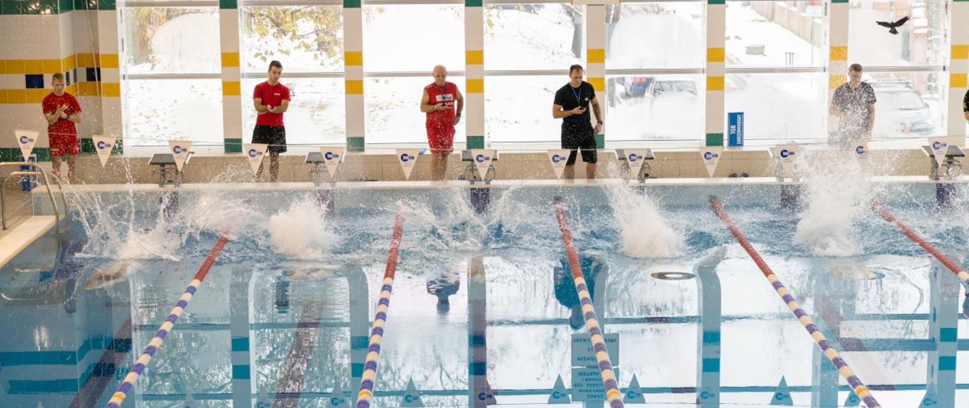 Zawodnicy, strażacy Państwowej Straży Pożarnej w strojach kąpielowych, po skoku do wody rywalizują w zawodach pływackich o puchar Opolskiego Komendanta Wojewódzkiego Państwowej Straży Pożarnej. W tle sędziowie zawodów.