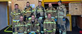 Grupa ośmiu strażaków z województwa opolskiego w pełnym umundurowaniu ochronnym pozuje podczas warsztatów doskonalących ROT asekuracyjnych RIT w Bazie Szkoleniowej JRG Knurów (20–21 stycznia 2026 r.). Przed nimi znajdują się torby ratownicze RIT i Cavitan, a tło stanowi ściana z elementami szkoleniowymi i grafiką maskotki strażackiej.