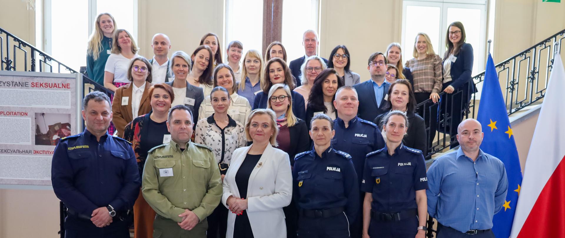 Grupowe zdjęcie uczestników oficjalnego spotkania, stojących na schodach wewnątrz budynku. W pierwszym rzędzie widoczne są osoby w mundurach polskich służb mundurowych (Policja, Straż Graniczna) oraz cywile. Obok grupy stoją flagi Unii Europejskiej oraz Polski.