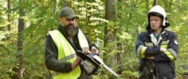 Zdjęcie wykonano na zrębie w lesie. W centrum widzimy obalonego świerka. Strażak z piłą oraz instruktor dokonują szkolenia z obsługi pilarki. 