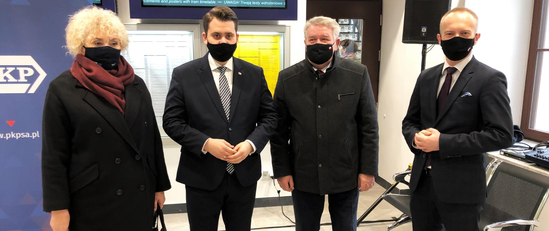 wyremontowana hala budynku dworca kolejowego w Mrozach. Na planie głównym cztery osoby. Jedna kobieta i trzech mężczyzn. Kobieta ubrana w ciemny płaszcz i bordową chustkę, obok mężczyzna ubrany w ciemny garnitur z krawatem w kratę, następnie mężczyzna ubrany w ciemny płaszcz i mężczyzna ubrany w garnitur. Wszyscy pozują do zdjęcia. 