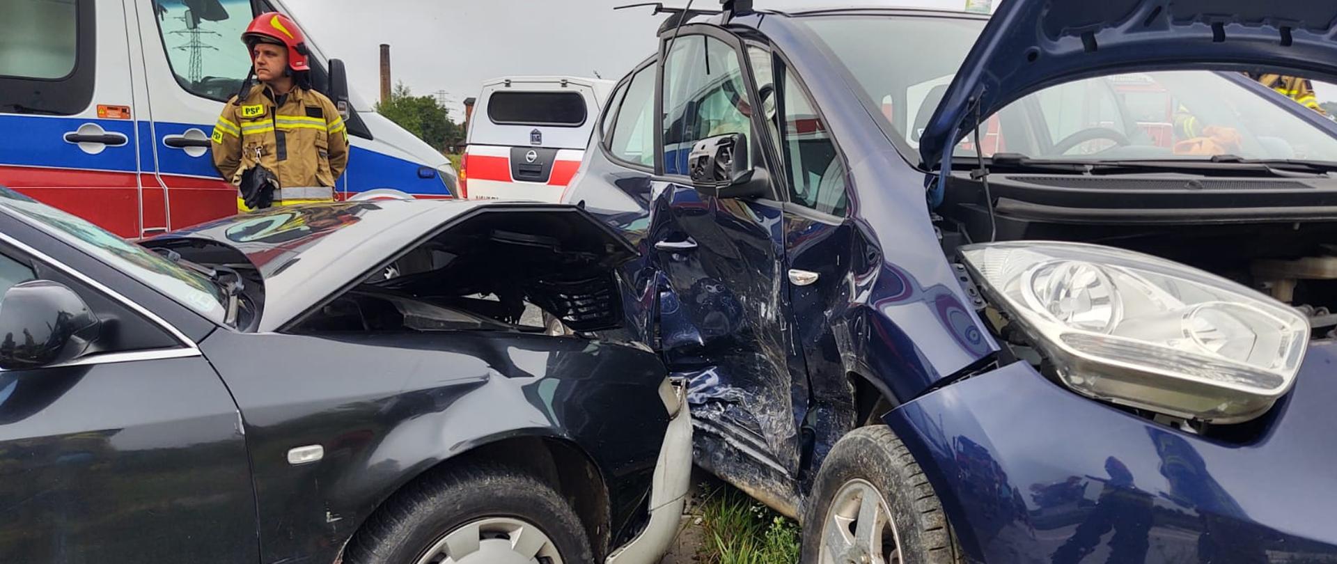 Widok samochodów, które uległy zderzeniu a w tle karetka, lekki samochód ratowniczy oraz strażak.