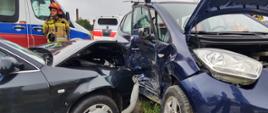Widok samochodów, które uległy zderzeniu a w tle karetka, lekki samochód ratowniczy oraz strażak.