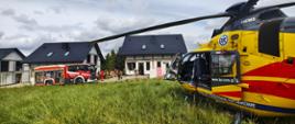 Śmigłowiec Lotniczego Pogotowia Ratunkowego stoi na trawie obok budowanych domów. W tle widoczny jest samochód straży pożarnej oraz grupa ratowników prowadzących działania przy jednym z budynków.