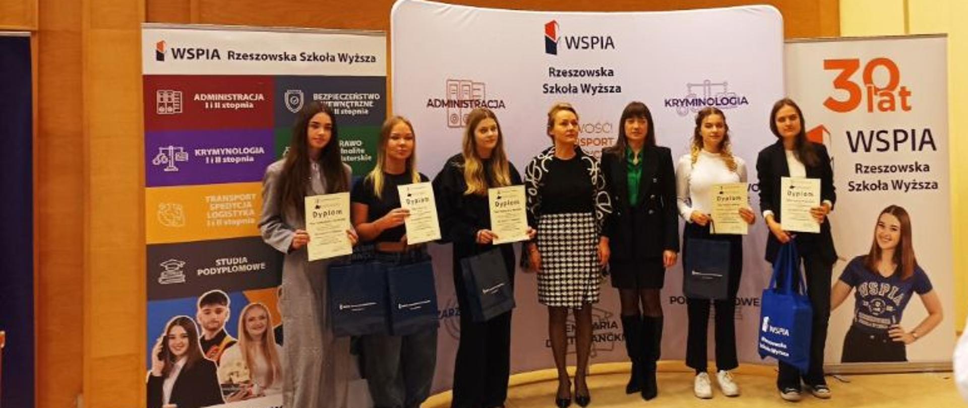 Uczestnicy konkursu stoją, w towarzystwie osoby wręczającej, z dyplomami w rękach i nagrodami rzeczowymi pod banerem informacyjnym WSPiA