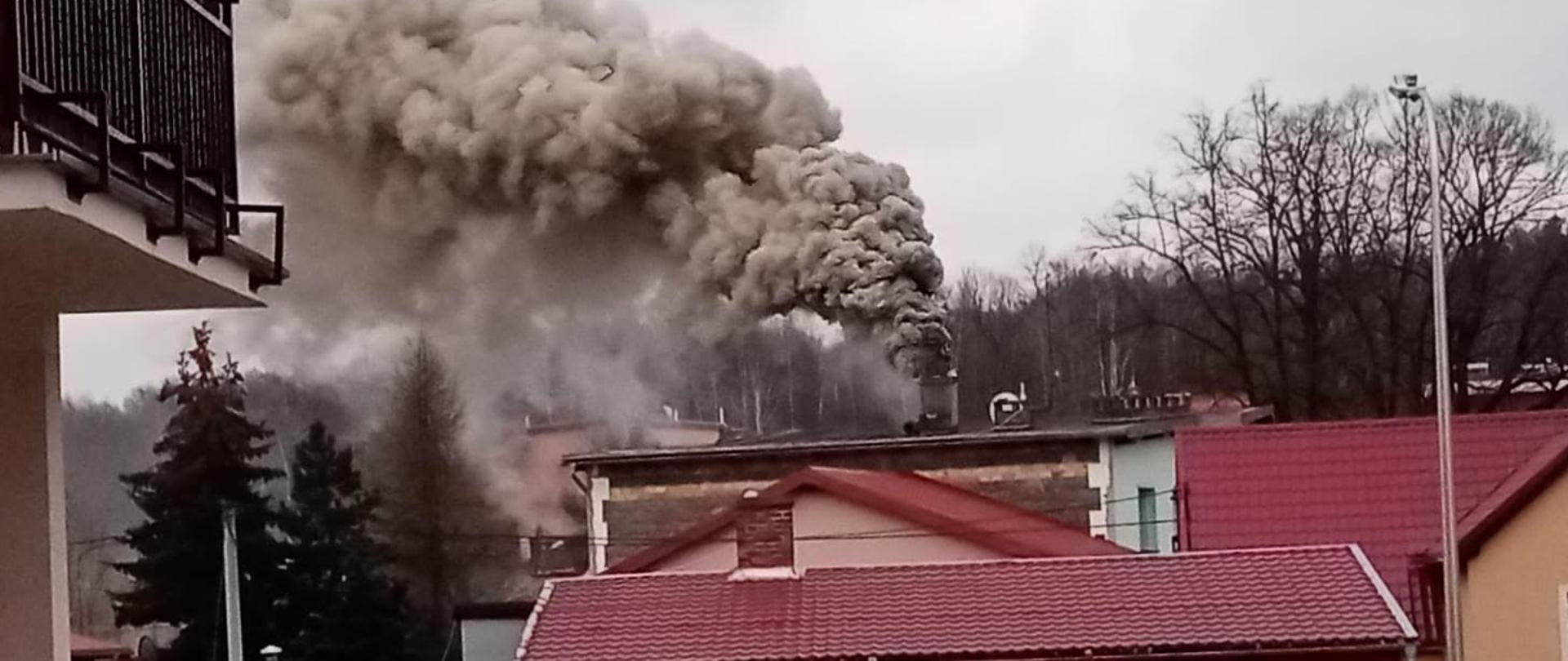 Na zdjęciu widać budynek koloru różowego. Nad budynkiem widać wydobywający się dym z komina. Dym jest gęsty i jest koloru biało szarego. Dym wydobywa się z komina. Dach budynku jest czerwony. 