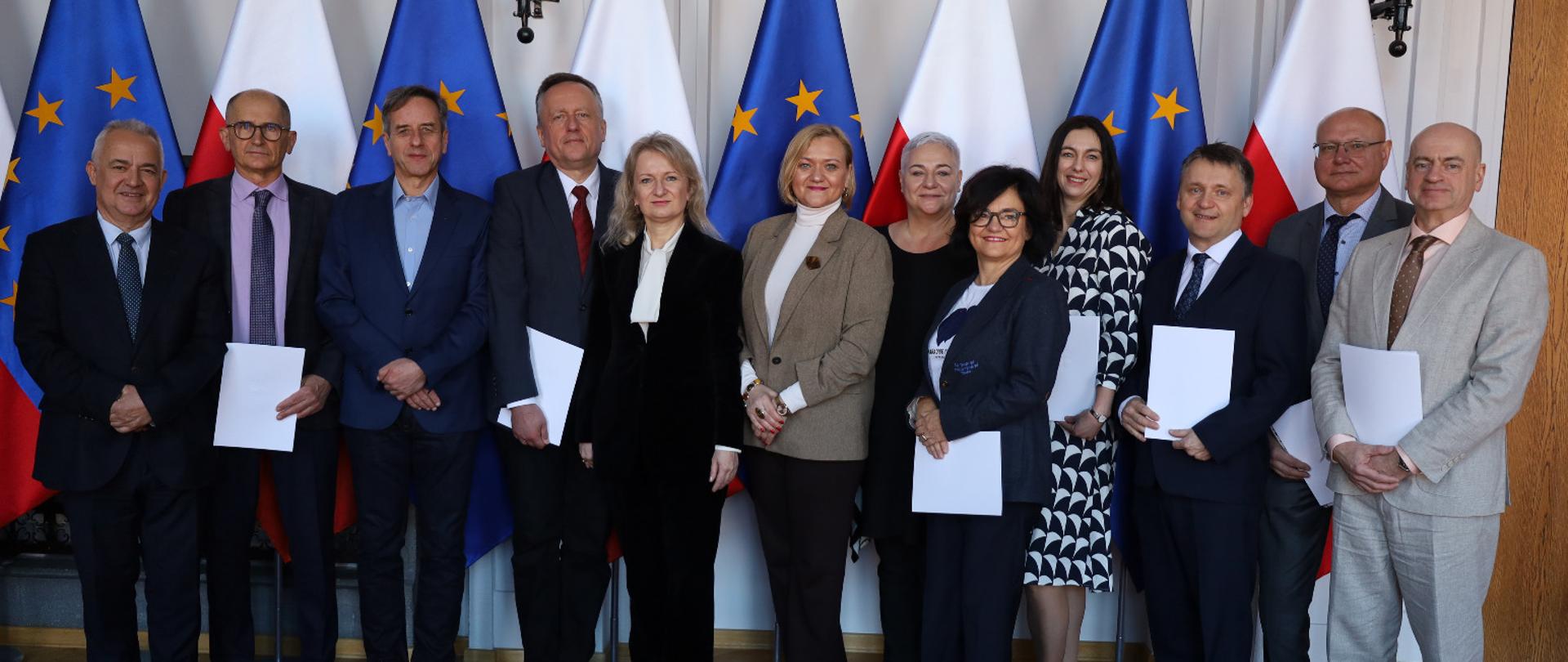kilkanaście osób stoi w sali i pozuje do zdjęcia na tle flag