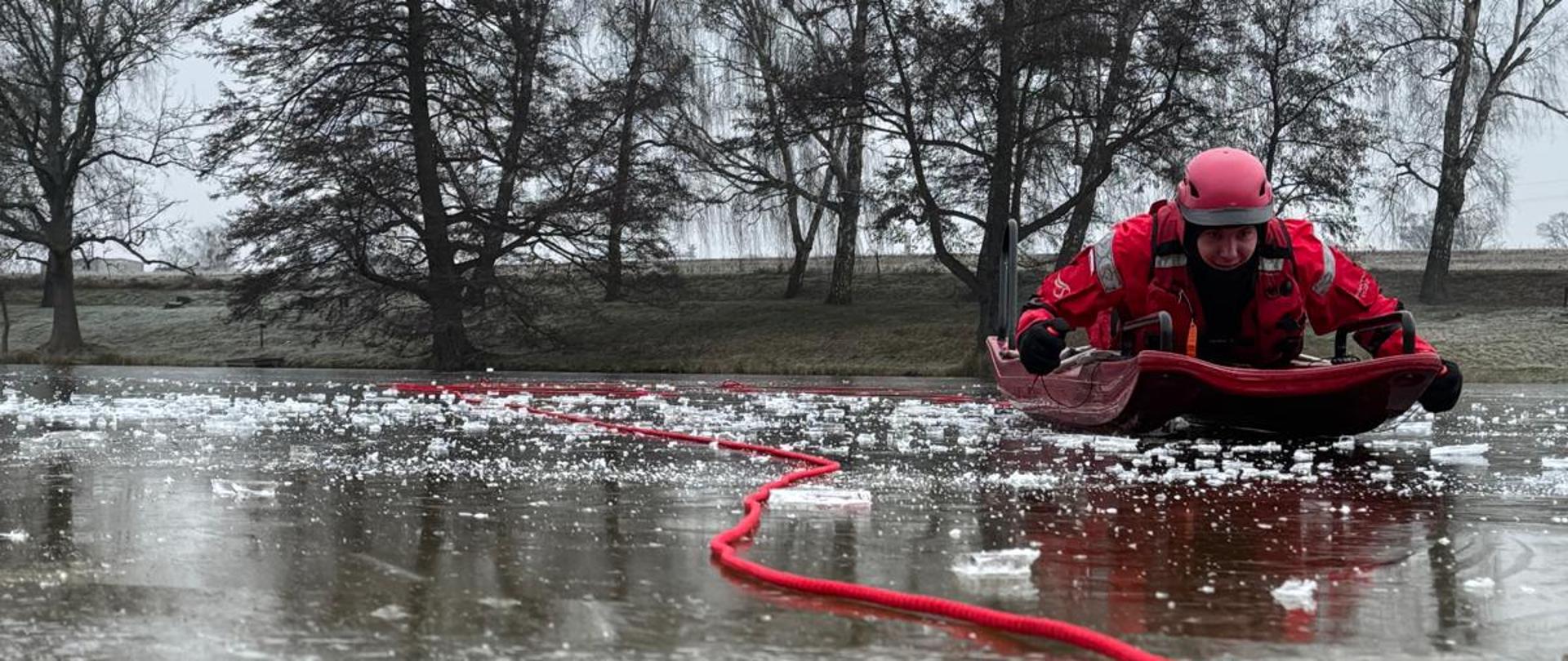Ujęcie przedstawia zbliżenie na ratownika poruszającego się po zamarzniętej tafli jeziora leżąc na saniach wodno-lodowych.