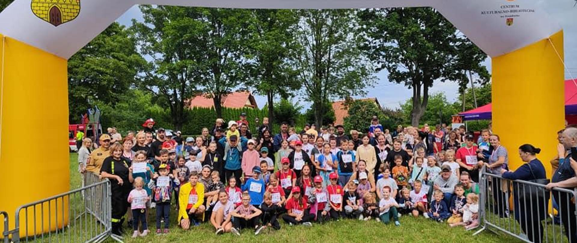 Na zielonej trawie boiska sportowego do zdjęcia pozuje duża grupa dzieci, uczestników i organizatorów biegu. dzieci kolorowo ubrane. duży żółto biały pompowany baner z napisem Frombork. W oddali zielone drzewa.