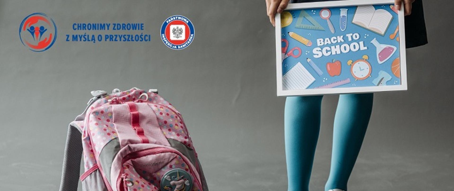 na zdjęciu widać nogi dziewczynki w białych sportowych butach i niebieskich rajstopach. W rękach powyżej kolan trzyma małą tablicę z napisem po angielsku "powrót do szkoły", obok dziecka na podłodze stoi różowy plecak szkolny. W lewym górnym roku logo Państwowej Inspekcji Sanitarnej "Chronimy zdrowie z myślą o przyszłości".