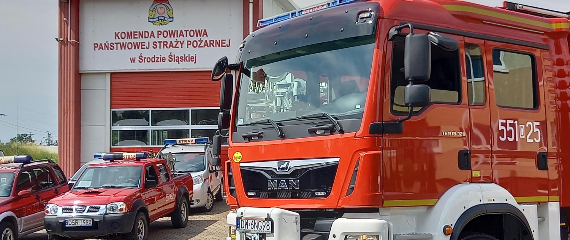 Samochód gaśniczy Man na tle innych samochodów strażackich i bramy garażowej z napisem Komenda powiatowa państwowej straży pożarnej w Środzie Śląskiej