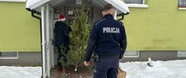 Policjant trzymający w prawej ręce torbę z ozdobami świątecznymi i pracownik MOPS z czapce Mikołaja z choinką w ręce wchodzą do klatki schodowej budynku wielorodzinnego. Na ziemi leży śnieg, w tle zielonkawa elewacja budynku i okna.
