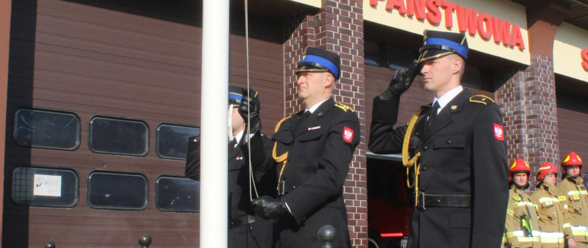 Trzyosobowy poczet flagowy strażaków w ubraniach galowych zawiesza flagę Polski na maszt przed budynkiem komendy.