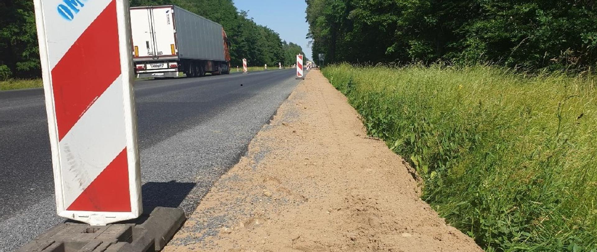 Zdjęcie wykonane z poziomu drogi. Z lewej strony widać drogę z nową nawierzchnią bitumiczną, z poboczem gruntowym. Na krawędzi, po obu stronach drogi, ustawione znaki biało-czerwone. Na drodze jadący samochód ciężarowy. Po prawej stronie zieleń przydrożna, a po obu stronach drzewa.