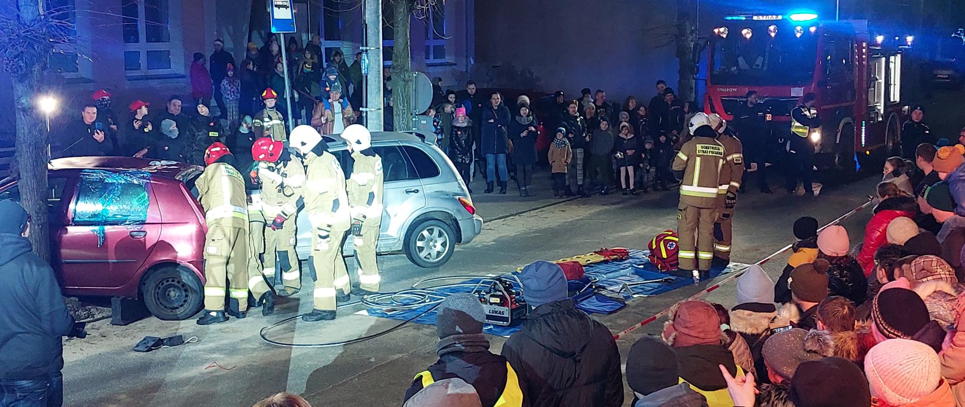 Strażacy wykonują pokaz ratownictwa technicznego. Dookoła zebrani zainteresowani obserwują wydarzenie. Zdjęcie wykonane w porze nocnej.