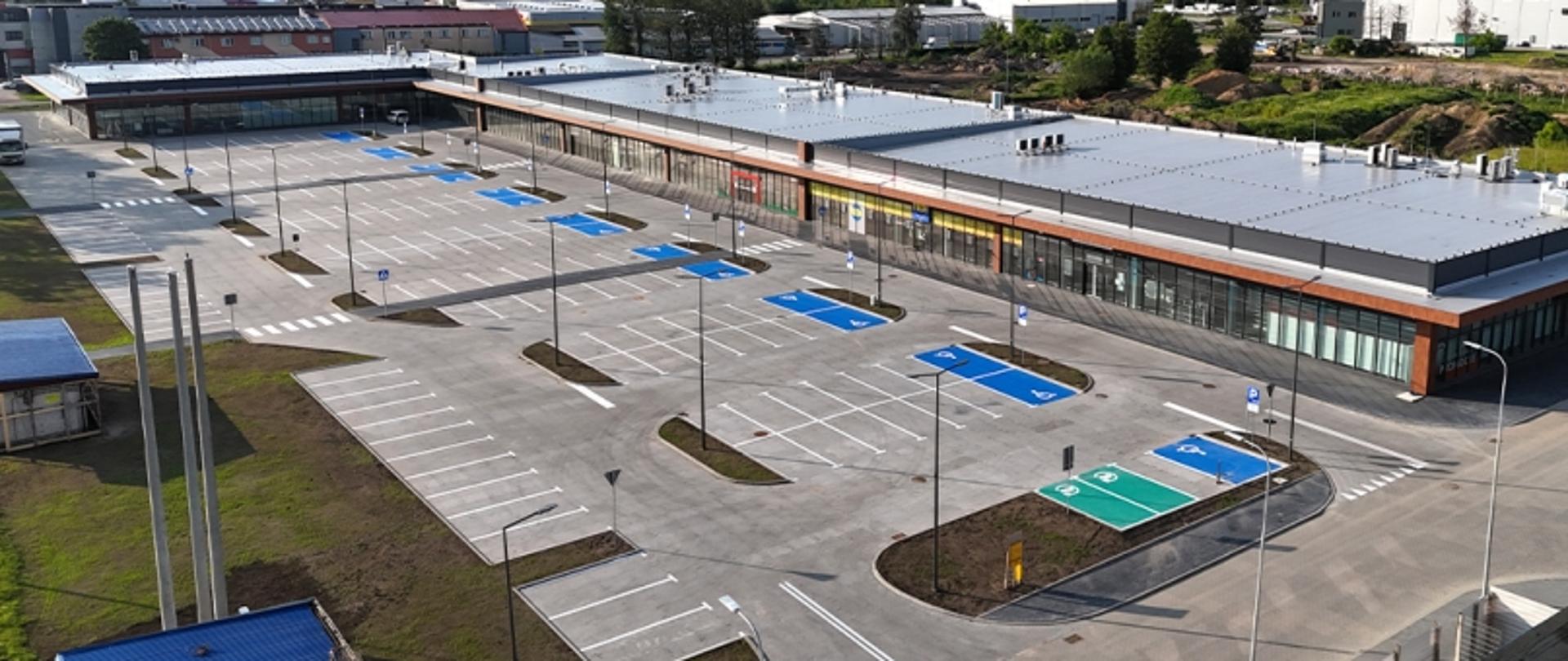 Na zdjęciu z lotu ptaka widać rozległy parking i parterowy, przeszkolony budynek z płaskim dachem.