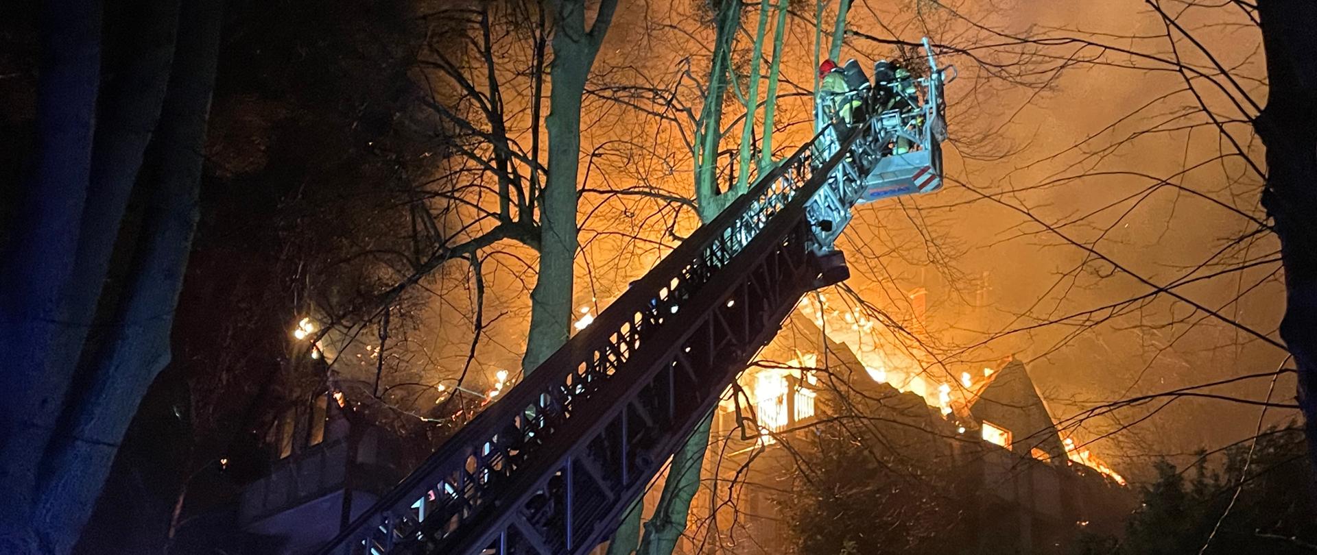 Strażacy w koszu drabiny mechanicznej przybliżają sie do płonacego budynku. W tyle widac płonącą ostatnią kondygnację