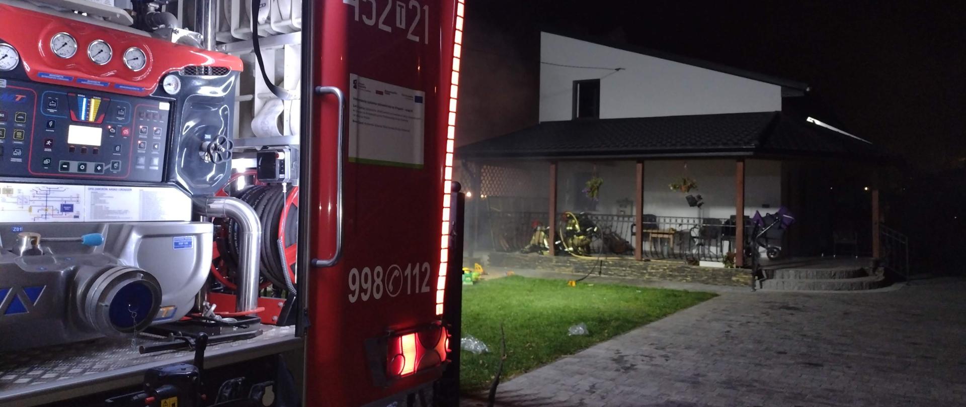 Na pierwszym planie widać tył samochodu gaśniczego w porze nocnej. W oddali dwóch strażaków wchodzących do zadymionej kotłowni.