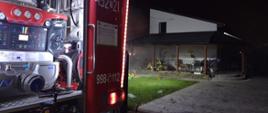 Na pierwszym planie widać tył samochodu gaśniczego w porze nocnej. W oddali dwóch strażaków wchodzących do zadymionej kotło