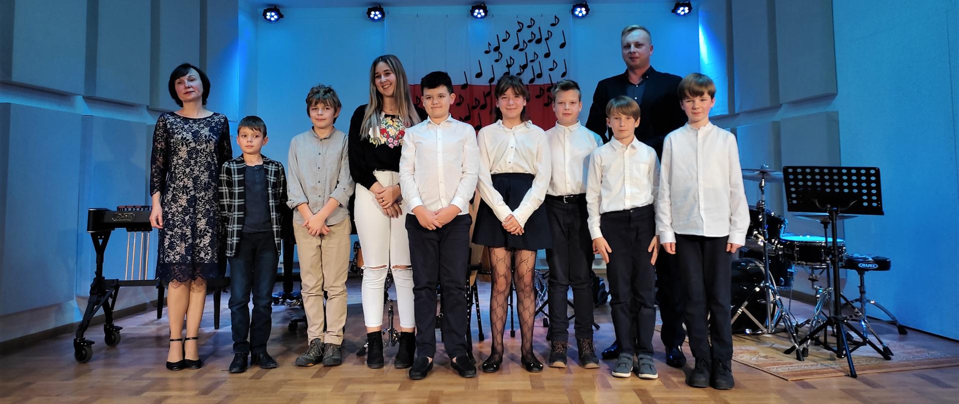 Zdjęcie przedstawia uczniów klasy perkusji wraz z nauczycielem oraz akompaniatorem po koncerice klasowym. Stoją na scenie szkoły, którą oświetla niebieskie światło. Po prawej stronie stoi perkusja. 