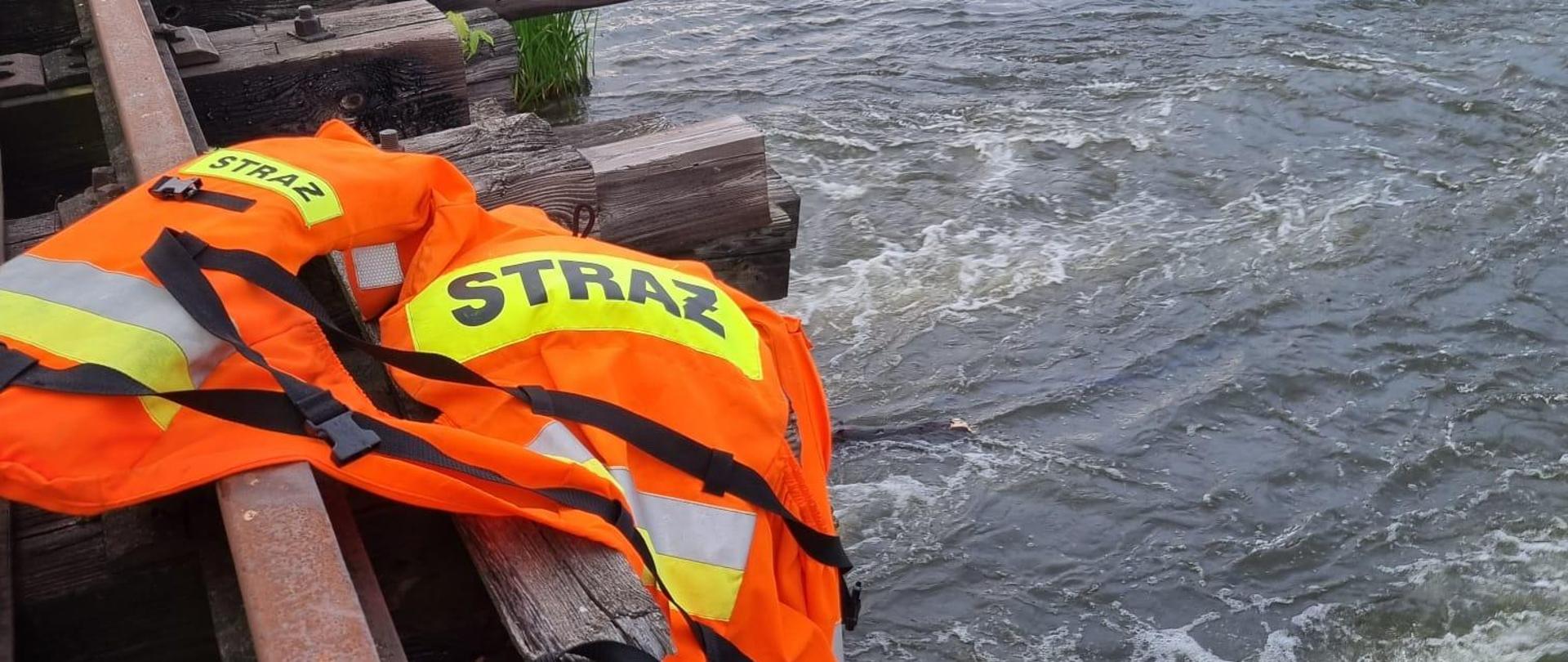 Zdjęcie na pierwszym planie przedstawia kamizelkę ratunkową w pomarańczowym kolorze. Na drugim planie widać zarośla oraz brzeg rzeki Nida.