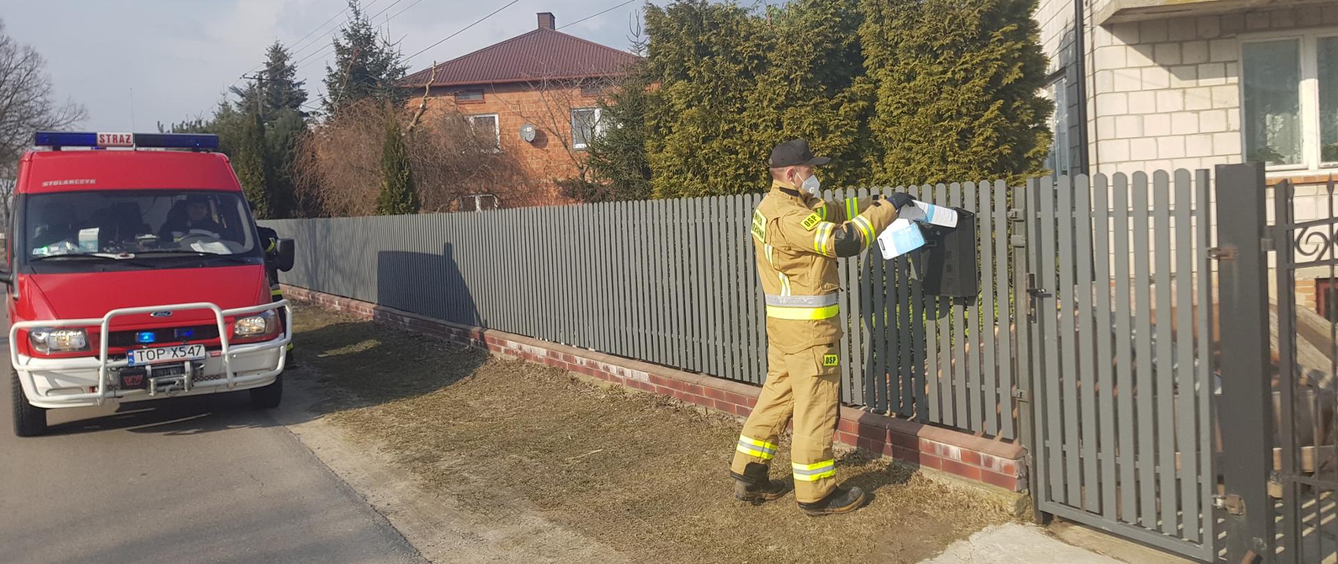 Zdjęcie przedstawia strażaka OSP wrzucającego ulotkę zawierającą informację o szczepieniach do skrzynki pocztowej. W tle widać zabudowania oraz czerwony samochód OSP.